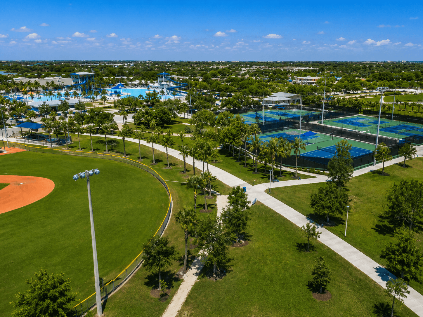 Aerial view of Pompano Beach Community Park showing green lawns, walking paths, palm trees, sports courts, a baseball field, and an aquatics complex under a clear Florida sky.